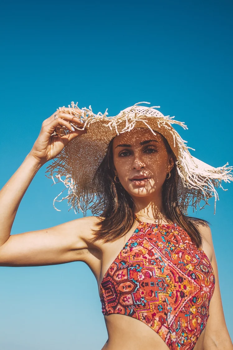 Kobieta w kolorowym kostiumie kąpielowym trzyma ręką słomkowy, duży kapelusz ochraniający głowę i twarz przed słońcem. Źródło: https://unsplash.com/